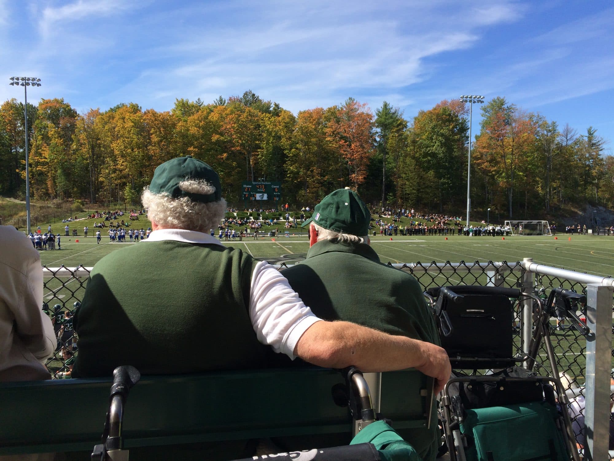 Dave Wolk and his father watching a game from the stands at Castleton's Spartan Stadium, fall foliage beyond.